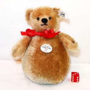 May include: A plush, golden-brown teddy bear with a red bow tie and a small red die with white dots. The bear has a round belly and a tag that reads "Steiff Rolly Polly Bär".