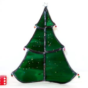 May include: A green stained glass Christmas tree ornament with dark metal soldering, featuring small colorful glass beads attached to the edges and a wire star at the top. A red six-sided die is placed to the left for scale reference.