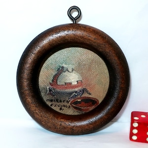 May include: A small, round painting in a dark wood frame with a metal loop for hanging. The painting depicts a still life with a white and red object and a bowl. The artist's signature "Shireen Eddings '81" is visible. A red die is next to the frame.