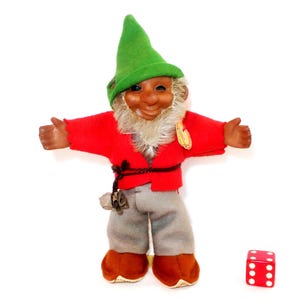 May include: A troll doll wearing a green felt hat, a red felt jacket, and grey felt trousers, with brown shoes and a white beard. A small brown pouch hangs from a dark cord around the waist. A red six-sided die is positioned to the right of the doll.