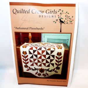 May include: A Quilted Crow Girls Designs pattern booklet titled Autumnal Pinwheels by Deirdre Bond-Abel. The cover features a photograph of a quilt with brown, orange, and cream pinwheel blocks, bordered by appliqued floral and leaf motifs.