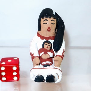 May include: A handcrafted ceramic figurine depicting a mother and child, with a red die next to it. The figurine features a woman in a white dress with red accents, holding a child. The die is red with white dots.