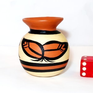 May include: A hand-painted ceramic vase with a round body and a flared rim. The vase features a cream-colored base with orange and black leaf designs. A red die with white dots is next to the vase.