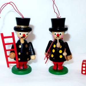 May include: Two wooden chimney sweep ornaments featuring black top hats, black jackets with gold dots, red trousers, and green circular bases. Each figure has a red nose, a small pipe, and a red hanging string. A red six-sided die sits nearby.