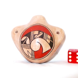 May include: A tan ceramic ocarina featuring a circular red and black geometric pattern on its front surface. The instrument has two small holes on the sides and is positioned next to a red six-sided die for a visual scale reference.