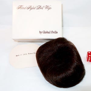 May include: A dark brown doll wig with a rounded shape, next to a white box with the text "Hand Styled Doll Wigs" and "by Global Dolls." A small, red die with white dots is also present. The wig is made of 100% human hair.