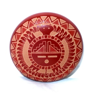 May include: A circular red object featuring a carved tan design of a central face with geometric patterns, including a stepped pyramid shape above the eyes and a sunburst-like border with triangular motifs surrounding the entire central figure.
