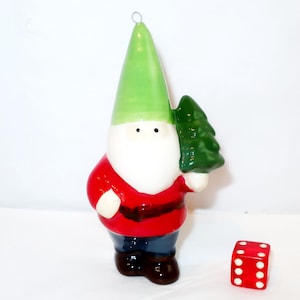 May include: A ceramic gnome ornament featuring a tall lime green hat, a white beard, a red jacket with a dark belt, and dark blue trousers. The gnome holds a small green pine tree in one hand. A red six-sided die is positioned next to the gnome.