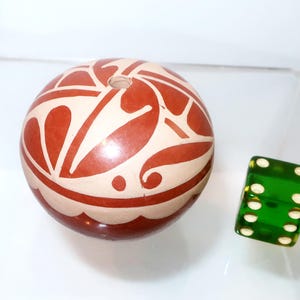 May include: A spherical ceramic object with a smooth, cream-coloured surface featuring a bold, dark red painted pattern of abstract leaf-like shapes and curved lines. A small circular opening is visible at the top. A green translucent die is nearby.