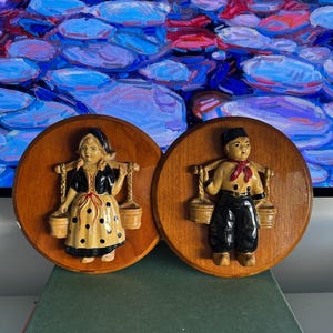 Puede incluir: Dos placas de pared de madera con esculturas en relieve de figuras holandesas. Las figuras visten trajes tradicionales y llevan cubos suspendidos de un yugo. Las placas son circulares y tienen un acabado marrón cálido.