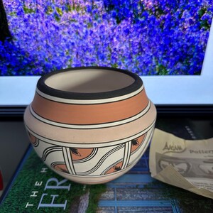 Peut inclure: Un bol en poterie fait à la main avec un intérieur crème, orné de bandes pêche, crème et noires. La partie inférieure présente un motif géométrique détaillé en noir et crème. Le bol est posé sur un fond floral flou.