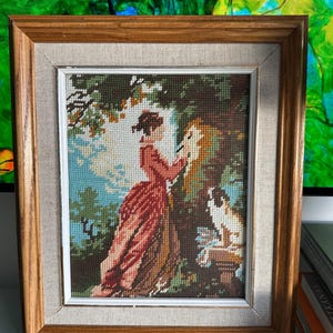 Puede incluir: Obra de arte de tapiz enmarcada que representa a una mujer con un vestido rojo y un perro en un jardín. La obra está enmarcada en un marco de madera marrón claro con un borde interior con textura de lino. La escena tiene un cielo azul y follaje verde.