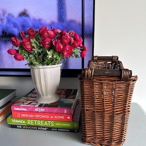 May include: A display of home decor featuring a white vase with pink flowers, placed on top of design books. A brown wicker basket with wooden handles is positioned beside the books, creating a warm, inviting atmosphere.