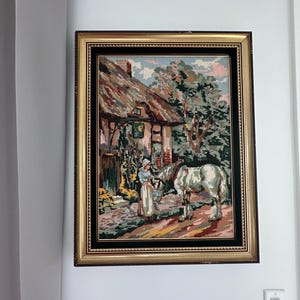 Puede incluir: Tapiz enmarcado que representa una escena rural con una cabaña con techo de paja, una mujer y un caballo blanco. La obra de arte presenta un marco dorado y un paspartú negro, añadiendo un toque de encanto vintage a la decoración de la pared.