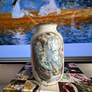 Puede incluir: Un jarrón de cerámica con un exterior texturizado blanquecino y un diseño pintado en tonos azules, marrones y verdes. El jarrón es cilíndrico con un borde ligeramente ensanchado. El fondo presenta una pintura de dos figuras en un bote.