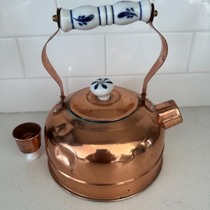 Puede incluir: Una tetera de cobre con un asa y un pomo de tapa con estampado floral blanco y azul. Una pequeña taza de cobre está al lado de la tetera. La tetera tiene un pico y un asa curva.
