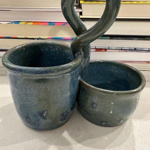 Puede incluir: Porta utensilios de cerámica hecho a mano en tonos azules y verdes. El soporte presenta dos cuencos conectados y un asa en forma de lazo. El esmalte tiene un aspecto moteado, lo que se suma a la estética única y artesanal. Un accesorio de cocina funcional y decorativo.
