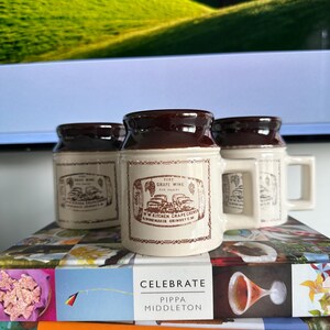 Puede incluir: Tres tazas de cerámica de color crema con bordes marrón oscuro y asas cuadradas. Cada taza presenta un estampado gráfico marrón de un barril con el texto PURE GRAPE WINE FOR SALE BY W.W. KITCHEN GRAPE GROWER & WINEMAKER GRIMSBY ONT.