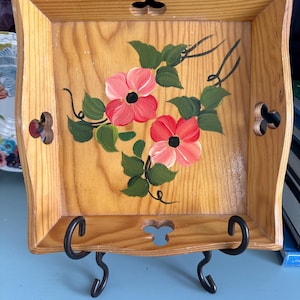 Puede incluir: Una bandeja cuadrada de madera con diseño floral. La bandeja marrón claro presenta un diseño pintado a mano de flores rosas y hojas verdes. Tiene recortes decorativos y se exhibe sobre un soporte de metal negro. Aproximadamente 25 cm cuadrados.