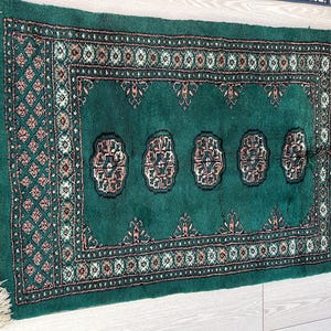 Puede incluir: Alfombra rectangular verde esmeralda con un patrón detallado. La alfombra presenta un diseño central de motivos florales repetidos en tonos marrón, crema y negro. El borde tiene un patrón geométrico y la alfombra tiene bordes con flecos.