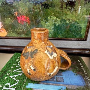 Puede incluir: Jarra de cerámica marrón con asa, decorada con figuras de bailarines. Las figuras son en negro, blanco y beige. La jarra está sobre un libro titulado "The Heart of France".