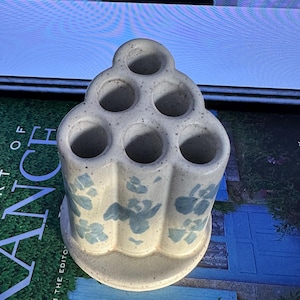 Puede incluir: Un portautensilios de cerámica con un acabado blanco roto moteado. Cuenta con seis aberturas circulares y está decorado con motivos florales azules. El soporte tiene una base redondeada y un diseño escalonado.