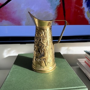 Puede incluir: Jarra de latón vintage con asa y boquilla. La jarra presenta un diseño en relieve de figuras y se exhibe sobre un libro verde. La jarra mide aproximadamente 15 cm de altura.