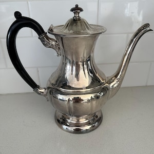 Puede incluir: Una tetera plateada con un asa negra y un pico curvo. La tetera tiene una tapa decorativa y un cuerpo redondeado. El asa es curva y el pico es largo y delgado. La tetera es probablemente de metal.