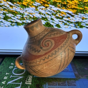 Könnte beinhalten: Eine kleine, handgefertigte Keramikvase mit rundem Körper und schmalem Hals. Die Vase hat eine beige Grundfarbe mit dekorativen Mustern in Rot, Schwarz und Braun. Sie hat zwei kleine Griffe und wird wahrscheinlich zu Dekorationszwecken verwendet.