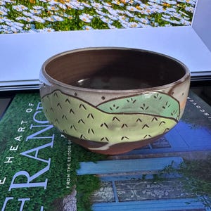 Puede incluir: Un cuenco de cerámica hecho a mano con interior marrón y exterior beige claro. El cuenco presenta un diseño de paisaje verde y marrón con pequeños detalles en negro. El cuenco está sobre un libro con las palabras "HEART OF FRANCE" visibles.