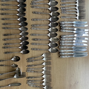 Puede incluir: Una colección de cubiertos antiguos, que incluye cucharas, tenedores y cuchillos, dispuestos sobre una superficie de madera. La cubertería tiene detalles ornamentales en el mango y una superficie pulida y reflectante. El juego incluye cucharas, tenedores y cuchillos de varios tamaños.