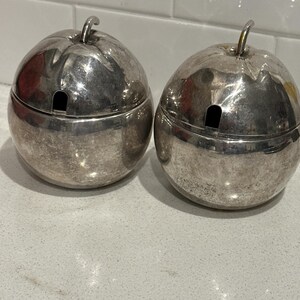 Puede incluir: Dos recipientes plateados con forma de manzana con tapas. Cada uno tiene un pequeño tallo y una ranura en la tapa. Los recipientes están sobre una superficie de color claro. Los recipientes parecen ser de metal.