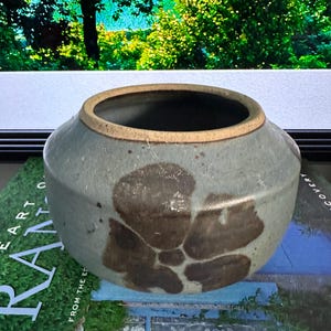 Puede incluir: Un pequeño jarrón redondo de cerámica con una abertura ancha. El jarrón es de color azul verdoso apagado con marcas de color marrón oscuro de forma orgánica. El borde es de color beige claro. El jarrón está sobre un libro con la palabra "ART".