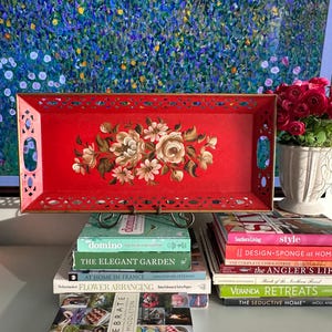 Puede incluir: Una bandeja rectangular roja con un diseño floral y bordes calados. Descansa sobre una pila de libros, incluyendo "The Elegant Garden" y "Celebrate". Un jarrón con rosas rojas es visible en el fondo.