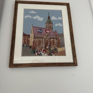 Puede incluir: Arte de punto de cruz enmarcado que representa una iglesia con un tejado estampado y un alto campanario. La obra de arte presenta una escena detallada con figuras con vestimenta tradicional, sobre un cielo azul con nubes. El marco es de color marrón oscuro.