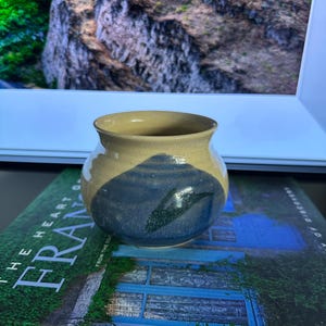 Puede incluir: Un pequeño jarrón de cerámica hecho a mano, de forma redondeada. El jarrón presenta una parte superior beige y una sección inferior azul con un diseño abstracto azul oscuro y verde. El jarrón está sobre un libro.