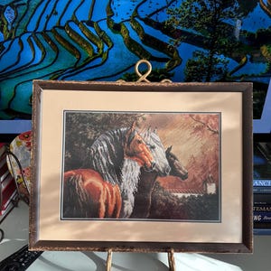 Puede incluir: Obra de arte en punto de cruz enmarcada que representa un grupo de caballos. Los caballos tienen pelajes marrones, grises y blancos. El fondo representa una escena rural con un edificio y árboles. El marco es de color marrón oscuro.