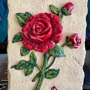 Puede incluir: Placa rectangular de piedra blanquecina con un relieve en relieve de una rosa roja y hojas verdes. La palabra "Rose" está escrita en escritura roja. La rosa tiene dos capullos. Objeto decorativo.