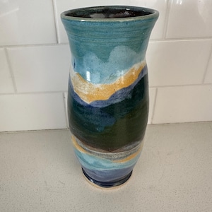 Puede incluir: Un jarrón de cerámica hecho a mano con la parte superior acampanada. El jarrón presenta un diseño de paisaje con tonos de azul, verde y amarillo. El jarrón mide aproximadamente 20 cm de alto y tiene un aspecto artístico único.