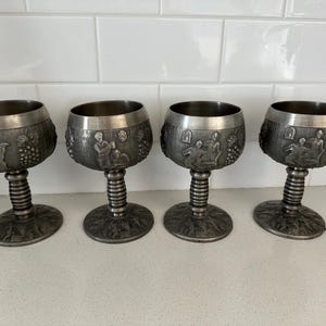 Puede incluir: Cuatro copas de color peltre con grabados en relieve detallados de figuras y vides. Cada copa tiene una base redonda, un tallo acanalado y un cuenco ancho. Las copas se exhiben sobre una superficie blanca.
