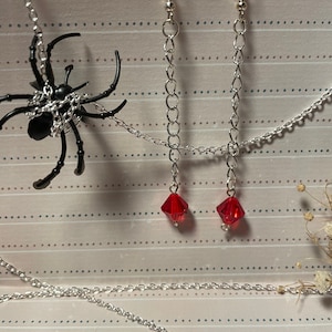 Könnte beinhalten: Ein schwarzer Spinnenanhänger mit einer silbernen Kette und ein Paar silberne Kettenohrringe mit roten Kristallakzenten. Der Schmuck wird auf einem gemusterten Hintergrund präsentiert, was auf ein gotisches oder Halloween-Thema hindeutet.