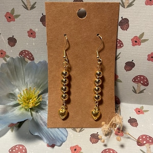 May include: Gold-colored dangle earrings. Each earring features a series of gold beads and a small heart charm at the bottom. The earrings are displayed on a brown card against a floral and mushroom-patterned background.