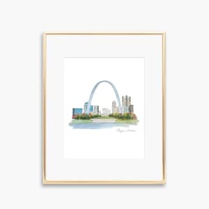 May include: A framed watercolor print of the St. Louis skyline, featuring the Gateway Arch and city buildings. The artwork is set against a white background and framed in a gold-colored frame. The print includes a river and floral accents.