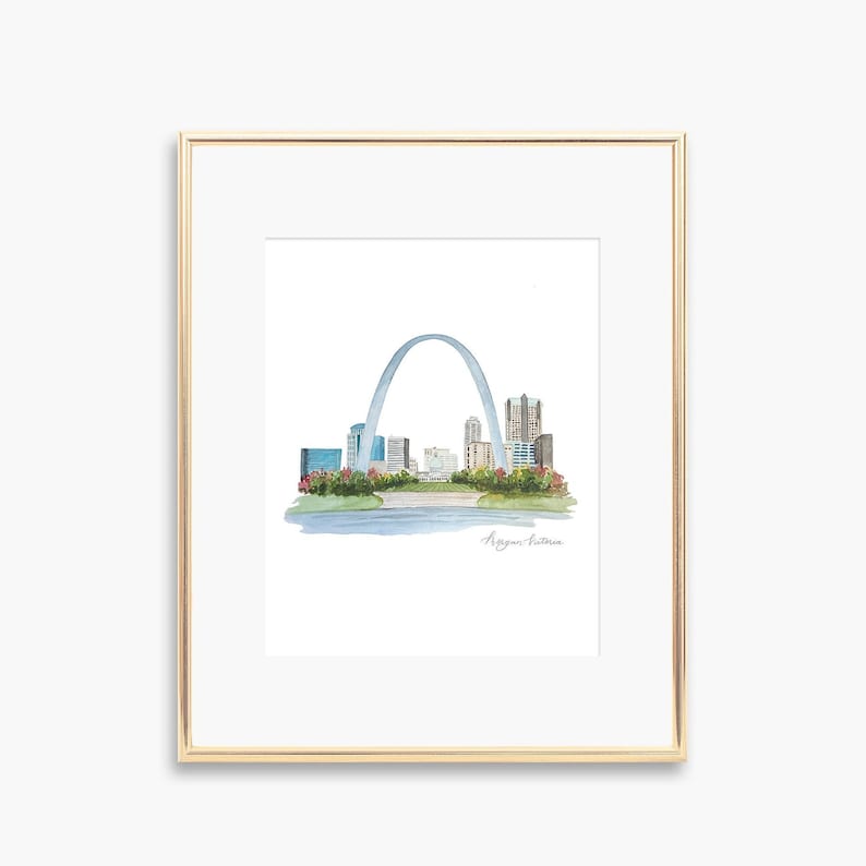 May include: A framed watercolor print of the St. Louis skyline, featuring the Gateway Arch and city buildings. The artwork is set against a white background and framed in a gold-colored frame. The print includes a river and floral accents.