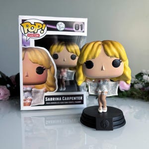 May include: A custom vinyl figure of Sabrina Carpenter, with a blonde hairstyle and a silver dress, stands on a black base. The figure is displayed in its original box, which features the Pop! Custom logo and the name "Sabrina Carpenter".