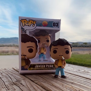 May include: A Funko Pop! vinyl figure of Javier Peña, a character from the TV series. The figure is in a clear display box with the name "Javier Peña" printed on the front. The figure is wearing a brown jacket, yellow shirt, blue jeans, and has a cigarette in his mouth.