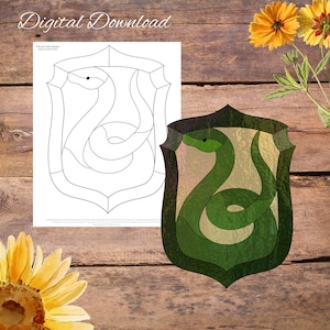 May include: A stained glass design of a green snake on a shield. The snake is curled up and has a green body with a yellow eye. The shield is green and has a black outline. The design is on a wooden background.