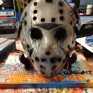 Pode incluir: Uma máscara de hóquei prateada com tiras pretas e um design preto e branco. A máscara tem vários orifícios e está riscada e gasta. A máscara está sentada em uma superfície colorida com a palavra "The Legend" escrita em preto.