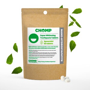May include: A brown paper bag of CHOMP Super Whitening Toothpaste Tablets, spearmint flavor. The label includes the text "Nano-Hydroxyapatite" and "60 tablets." Several mint leaves and three white tablets are scattered around the bag.