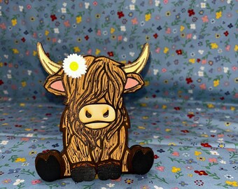 Puede incluir: Figura de madera de una vaca de las Tierras Altas con una margarita en la cabeza. La vaca es marrón con pezuñas y cuernos negros. El fondo es un estampado floral azul.